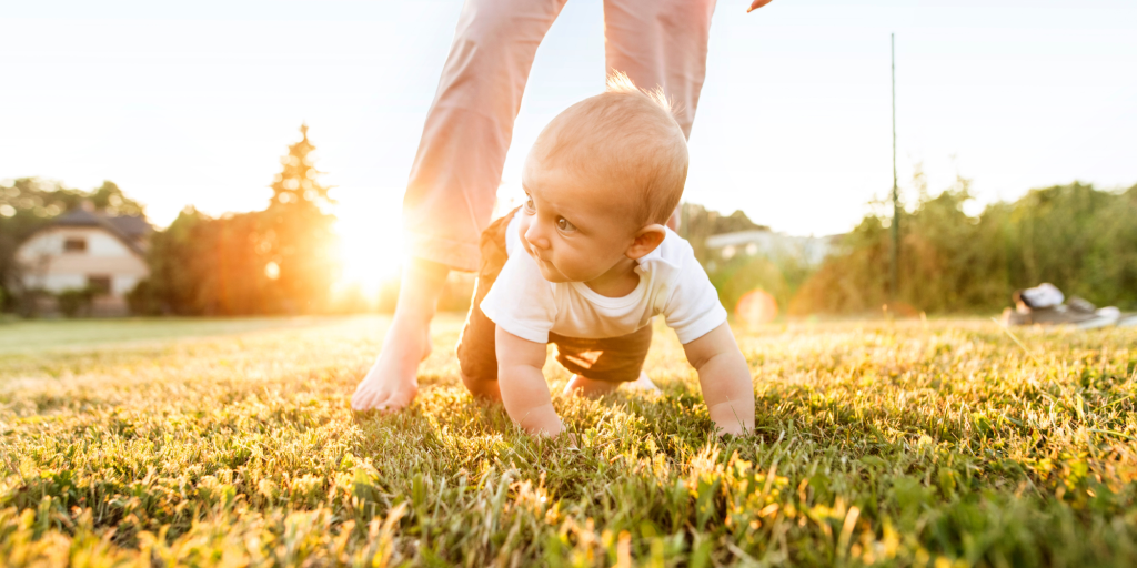 A baby crawls along the grass while playing outside with an adult