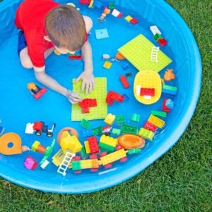 A child in a red swim shirt sits in a blue kiddie pool. The pool has water and LEGO Duplo bricks in it.