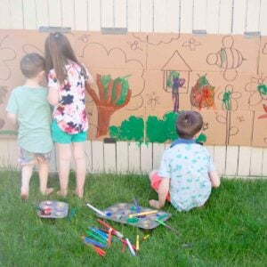 Three kids sit at the base of a fence painting a large hand drawn coloring page.
