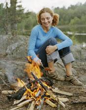 A woman cooking a hotdog on a stick over a campfire