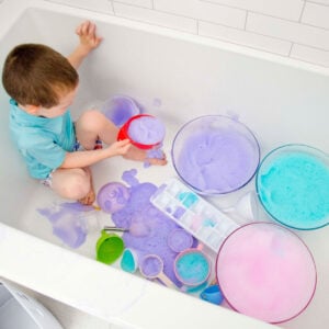 A child in a bathing suit sits in a bathtub. There are bowls of purple, blue, and pink bubble foam around him.