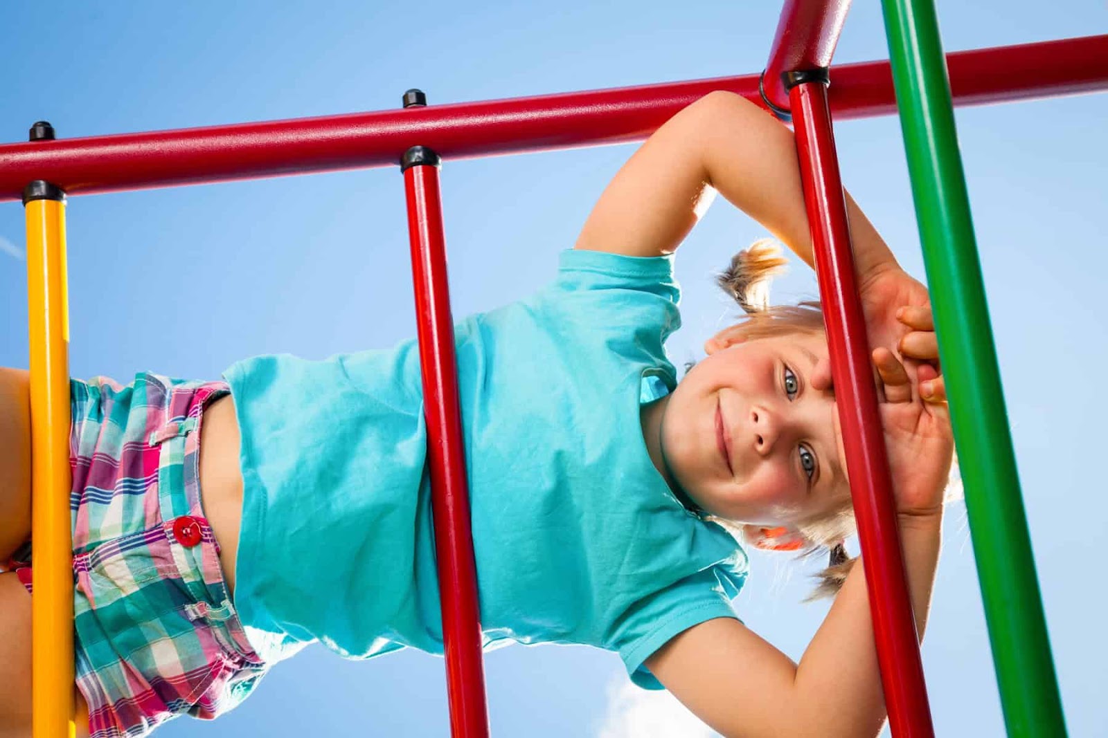 young girl with pigtails, wearing a turquoise shirt and plaid shorts laying across the red monkey bars