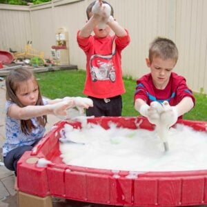 Three kids at a water table outside. The table is full of bubble foam. The kids are covered in it.