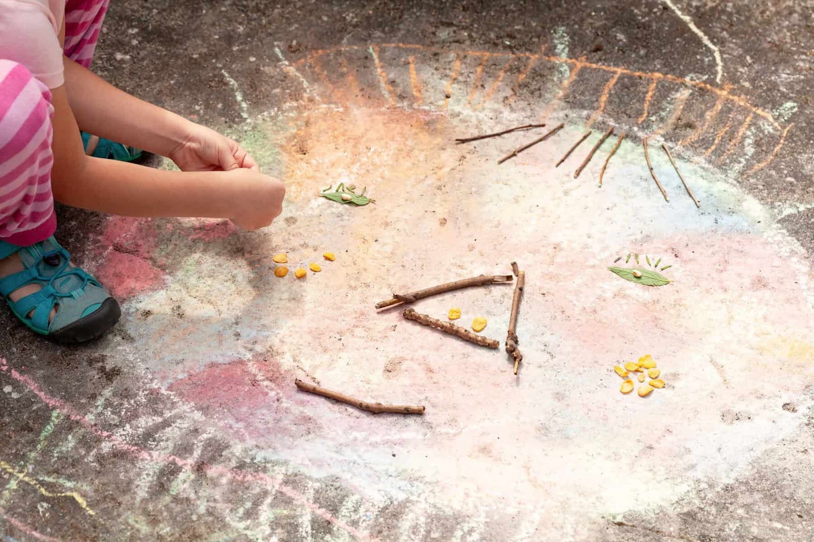 a giant chalk face with small sticks for a nose and mouth. small leaves for the eys and eylashes. you see the hands and feet of a child creating the art