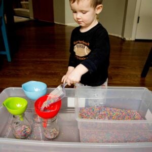 A child scoops rainbow rice into a red funnel. He is 2 years old and wearing pajamas.