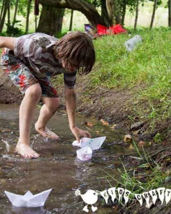 A child in a stream bends to reach a paper boat.