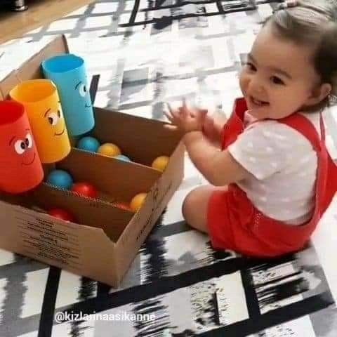 This may contain: a baby sitting on the floor next to a cardboard box with cups in it and smiling