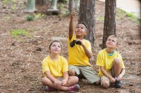 Three elementary school children kneeling on the ground in a forest. One is holding a pair of binoculars and pointing upward toward the trees. The other two are looking up to where he's pointing.