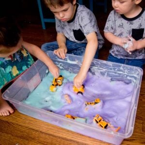 Three kids sit around a sensory bin filled with turquoise and purple bubble foam. They are pushing trucks through the foam.