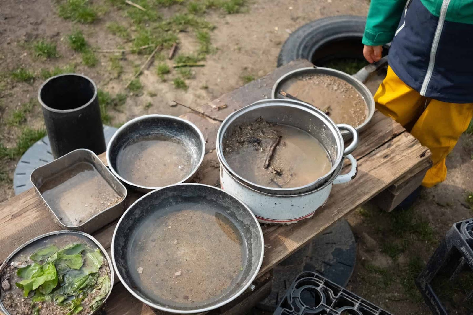 an outdoor mud kitchen with a young child off to the side wearing bright yellow pants and a blue and green jacket